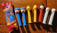 Photo of 8 Pez dispensers featuring Peanuts characters on a wooden surface. One dispenser is unopened in packaging, others are opened and arranged in a row by color and character.