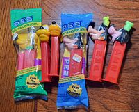 Six Disney PEZ dispensers arranged on a wood surface with two in original sealed packaging and four opened. Four are Pluto character heads and two are Goofy character heads.