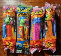 Photo of four unopened Pez Sourz candy dispensers with different colorful character heads in original sealed packages showing some leakage contained inside.