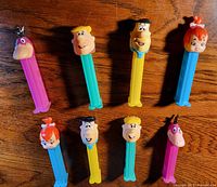 Photo showing all 8 Flintstones PEZ dispensers arranged on wood surface. Characters visible include Dino, Fred Flintstone, Barney Rubble, Wilma Flintstone, Betty Rubble, Pebbles Flintstone.