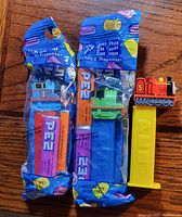 Photo showcasing three PEZ Thomas The Tank Engine candy dispensers; two are unopened in their original blue packaging with candy sticks inside, one opened dispenser is yellow with a red engine figure on top.