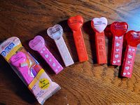Seven Pez Valentine's Day heart-themed dispensers displayed on a wooden surface; one dispenser is unopened and packaged.