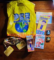 Yellow PEZ Visitor Center plastic bag, lanyards with passes, boxed PEZ candy dispenser, assorted postcards and pamphlets, and two commemorative visitor center pins arranged on wooden surface
