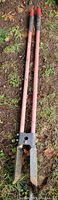 Full view of the post hole digger showing the two wooden handles with red tips and the metal digging blades at the base laid on the ground.