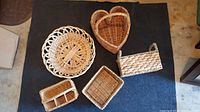 Five wicker baskets of various shapes and sizes displayed on dark fabric with floor background. Visible are round, heart, rectangular with handles, square, and divided compartment baskets arranged in rows.