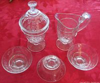 Full set of five clear glass items photographed on red cloth background, showing covered jar, creamer pitcher, and three dessert dishes.