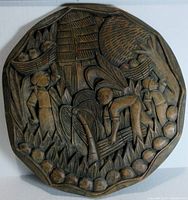 Full view of the round, octagonal hand carved wooden plaque showing the village scene with three figures and huts.