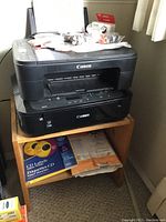 Two Canon printers stacked on a small wooden shelf alongside printer paper and CD labels below.