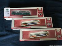 Three Corgi boxes showing green-cream livery box on top and two white multi-color livery boxes below