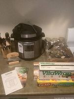 Wide view showing Cosori pressure cooker, wooden knife block with knives, assorted cutlery tools in plastic bags, jars with lids, and multiple boxes of food wrap (VitaWrap and Glad). Manual and recipe booklet also visible.