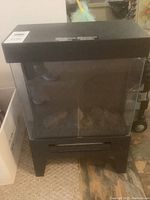 Front view of black electric fireplace showing glass doors, vent panel, and internal artificial logs.