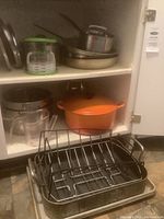 Overview of cabinet with several pots, pans including orange Le Creuset Dutch oven, metal roasting pan with rack, measuring cup, stainless steel sauce pot and frying pans, and a green and white plastic Starfrit burger maker