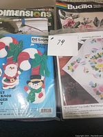 Photo showing unopened Dimensions felt Christmas decoration kit with Santa and snowman, Bucilla needlepoint pillow kit with floral design, and Bucilla stamped applique kit for Rose Percale quilt
