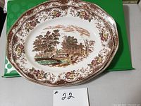 Oval platter with scenic image of people resting near trees and water, surrounded by floral border.