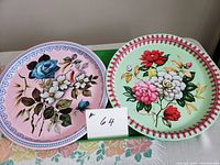 Two round metal trays side by side on a floral tablecloth. One tray is pink with blue rose and white flowers and blue border; the other is green with mixed colored flowers and red border.
