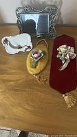 Full lot displayed on wooden table showing silver-tone ornate picture frame, two ceramic flower wall plaques with velvet backings, porcelain dish with flower decoration.