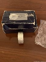 Single textured silver-tone napkin ring next to the original worn box and plastic wrapping on wood surface.