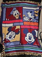 Full view of the blanket showing Disney characters Mickey Mouse, Minnie Mouse, Donald Duck, and Goofy, with colorful stripes and fringed edges.