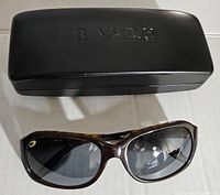Top view of closed hard case with embossed Ralph Lauren logo and sunglasses laid in front showing front lens and frame shape.
