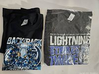 Two black Tampa Bay Lightning Stanley Cup Championship T-shirts side by side, one folded in packaging, one loose with graphic showing players and Stanley Cup trophy.