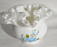 View of the vase showing white opaque glass with hand painted blue roses and leaves, and the wavy clear glass rim.