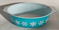 Front angle view of turquoise Pyrex casserole dish decorated with white snowflakes around the side and light aqua interior. No lid included.