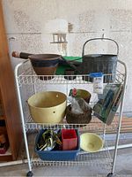 Photo of white metal rolling cart with three shelves, holding garden tools and pots, including a watering can, pots, gloves, small basket, seed kits and plastic containers.