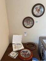 Three vintage collectible plates on a wooden surface. Two plates are standing facing the wall mounted on the wall, one plate is lying on the table with documentation and wooden frames next to it.