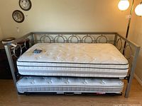 Metal framed daybed with circular patterned side panels and two white tufted mattresses, one on the daybed and one on the trundle underneath, placed against a wall with floor lamp and hardwood flooring.