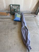 Patio umbrella with metal pole wrapped in gray cover, green metal umbrella stand, and sealed blue package of outdoor furniture covers arranged on concrete floor inside garage.