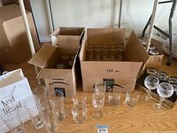 Multiple glassware items including tall and short glasses arranged on a wooden floor along with several boxes containing more glasses.