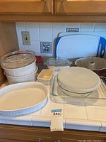 Full set of kitchenware on counter including CorningWare cake pans, glass mixing bowl with lid, casserole dish, Bundt pan, and cutting boards.