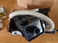 Oreck XL handheld vacuum cleaner packed in cardboard box with black body, white flexible hose and black attachments visible.