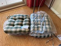 Four green and beige plaid seat cushions, two visible fully and two stacked in plastic wrapping.