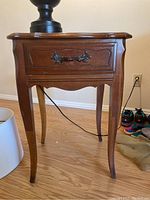 Front view of vintage wooden side table showing single drawer with carved decorative panel and metal handle, slender legs and scalloped apron.