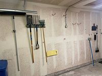 Wall-mounted gardening tools including rakes, shovel, and broom handle mounted on a garage wall with hooks.