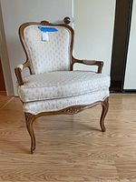 Front view of the vintage carved wooden armchair with beige upholstery, showing carved floral crest and padded seat, arms, and legs.