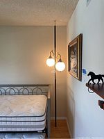 Full view of the vintage floor lamp wedged between floor and ceiling next to a bed frame and painting. The lamp has a tall black pole and two lit white globe light shades with bronze metal holders.