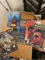 Open box showing a stack of over 100 sealed, bagged, and boarded comics. Top comics visible include Hellboy Winter Special, Mad Max Fury Road, Negative Space, Mirrors Edge, and Pacific Rim The Rift 1.