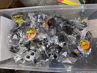 Box filled with individually bagged enamel pins, visible designs include pumpkins, skulls, and masks or robot faces, showing variety in the lot.
