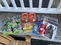 Wide shot showing entire lot of assorted collector toys displayed on stone surface outside near white railing including Scatter Brainz packs, Tina Belcher Sodaz boxes, Adventure Time bath set, Raving Rabbids figure, Star Wars figure and poster.