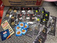 Photo shows 29 enamel collector pins laid out on carpet. Pins include Marvel branded pins, comic style 'POW' pins, Canadian Olympic pins with maple leaf and sports motifs, assorted gold tone pins, and other cultural symbols.