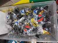 Clear plastic bin filled with multiple individually bagged enamel pins featuring Godzilla and other characters, showing several distinct designs including Godzilla's red and black logo and blue skull pins.