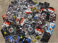 Photo showing all 65 enamel pins spread out on a flat surface, featuring many different designs including cartoon characters, logos, and symbols. Some pins are packaged on backing cards, others in plastic wrapping.