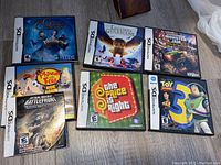 Seven Nintendo DS video games in cases, arranged in two rows on a carpeted floor. Titles visible include The Golden Compass (sealed), Legend of the Guardians, Monster Jam Urban Assault, Phineas & Ferb Ride Again (case/manual only), Star Wars Battlefront, The Price is Right, and Toy Story 3.