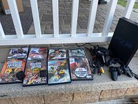 Photo of Sony PS2 console, controllers, cables, and eight video game cases displayed on outdoor concrete surface.