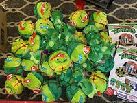 Photo showing about 22 Teenage Mutant Ninja Turtle plush toys, 12 green Cthulhu plush toys, and two boxed Teenage Mutant Ninja Turtle collectible items on a carpeted floor.