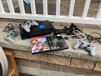 Lot shows PlayStation 2 slim console with controllers, cables, memory card, and game discs on a stone surface outside.