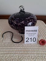 Purple blown glass hummingbird feeder displayed with black hanging hook and red flower feeder attachment, shown on quilted surface.