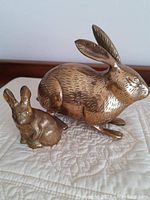 Two solid brass rabbit figurines placed on a quilted surface. Larger squatting one on the right, smaller standing one on the left, showing detailed etched fur and polished finish.